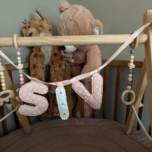 Op de afbeelding: Een houten babygym met hangende houten kralen in pasteltinten en een stoffen slinger die de naam SIV spelt. De letters zijn gemaakt van lichtroze stof met witte stippen en een witte middelste letter met een bloemenpatroon.