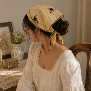 May include: A pale yellow triangular headscarf featuring black floral embroidery and a decorative black scalloped edge. The fabric has a subtle textured finish, and the scarf is tied at the back of the head with two pointed ends hanging down.