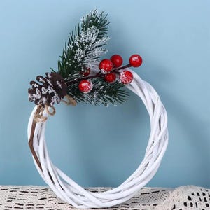 May include: A circular white woven wreath decorated with a cluster of green pine needles dusted with artificial snow, a brown pinecone, and several bright red berries. The decorative arrangement is attached to the top of the white twig frame.