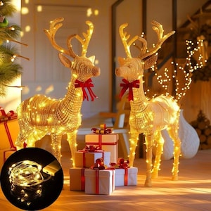 May include: Two standing reindeer figures constructed from gold-toned wire mesh, illuminated by warm white LED string lights. Each reindeer features a red ribbon bow around its neck. A coil of copper wire LED string lights is shown in a circular inset.