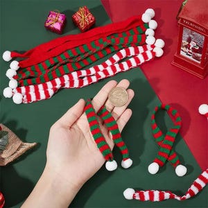 May include: A collection of miniature knitted scarves featuring red and green or red and white horizontal stripes, each finished with small white pom-poms at the ends. A gold-coloured coin is held to show scale alongside the small festive accessories.