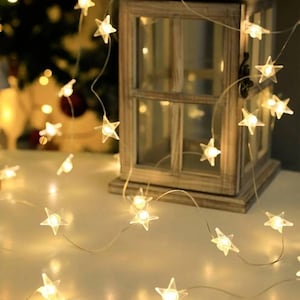 Könnte beinhalten: Eine Lichterkette mit sternförmigen LEDs, die ein warmweißes Licht ausstrahlen. Die Lichter sind an einem feinen, dünnen Draht befestigt, der um einen hölzernen Laternenrahmen drapiert ist. Jeder kleine Stern enthält eine helle Glühbirne.