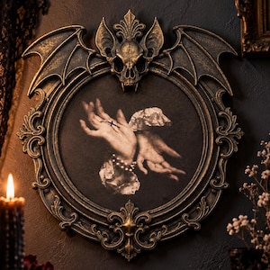 May include: An oval wall frame with a dark bronze finish, featuring a sculpted bat skull at the top and ornate scrollwork. The central artwork depicts two pale hands with a small cross and a pearl bracelet against a dark, textured background.