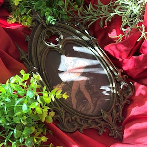 May include: An oval-shaped, dark bronze-toned picture frame with intricate gothic-inspired architectural detailing, including pointed arches and fleur-de-lis motifs. The frame contains a black and white illustration of a person in historical attire.