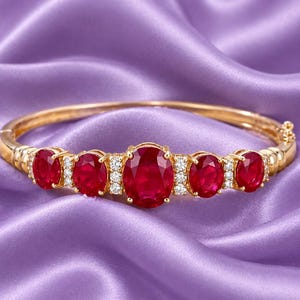 Puede incluir: Una pulsera rígida de oro con bisagra que presenta cinco rubíes rojos de corte ovalado dispuestos en fila, siendo el rubí central el más grande. Pequeñas piedras redondas y transparentes están engastadas entre cada rubí, dando brillo.