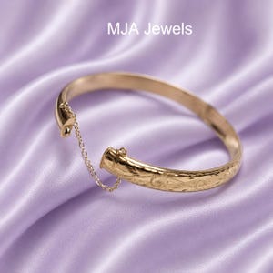 May include: A gold-toned hinged bangle bracelet featuring an intricate engraved scroll pattern and a delicate safety chain connecting the two ends. The text MJA Jewels is visible at the top of the frame. The bracelet has a polished finish.