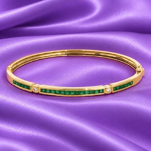 Op de afbeelding: Een scharnierende armband van gepolijst geelgoud met een kanaalgezette rij vierkante groene smaragden, geaccentueerd door twee kleine ronde, heldere diamanten. De armband is voorzien van een veilig sluitmechanisme aan de zijkant.