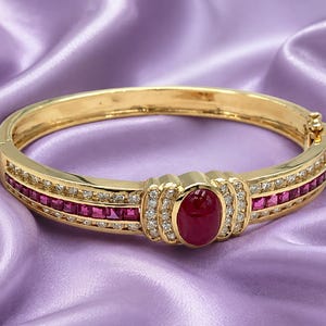 May include: A gold hinged bangle bracelet featuring a central oval cabochon ruby surrounded by small round diamonds. The band is accented with a row of square-cut rubies flanked by two rows of small round diamonds on each side of the center.