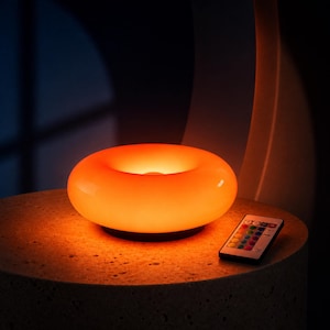May include: A round, donut-shaped LED lamp emitting a warm orange glow, featuring a smooth, translucent surface. Beside the lamp sits a small, rectangular remote control with multiple colored buttons for adjusting light settings and color modes.