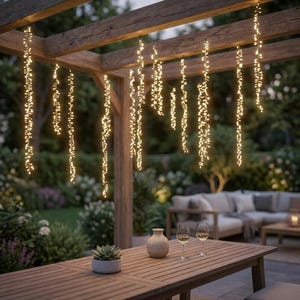 May include: A string of warm white LED fairy lights hanging vertically from a wooden pergola structure. The lights are arranged in multiple strands, creating a glowing curtain effect above a wooden outdoor table with a small succulent plant.