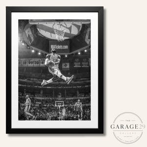 May include: A black and white framed photograph depicting a basketball player wearing a jersey with the number 23 performing a slam dunk in a crowded arena. The image features a basketball hoop, stadium lights, and the text NBATickets.com visible.