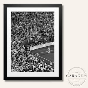 May include: A black and white framed photograph depicting a high-angle view of a soccer stadium. A lone player stands near the corner flag on the pitch, while a dense crowd of spectators fills the stands, creating a stark contrast in the scene.