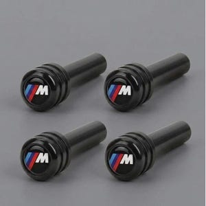 Op de afbeelding: Een set van vier zwarte cilindrische ventieldopjes, elk voorzien van een rond embleem met de letter M in het wit en een driekleurige streep in blauw, paars en rood op een zwarte achtergrond, ontworpen voor autobandenventielen.