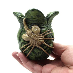 May include: A small, dark green, textured egg-shaped sculpture featuring a tan, multi-legged creature attached to the front. The object has a rough, organic surface with several pointed, leaf-like flaps at the top opening, resembling a biological pod.