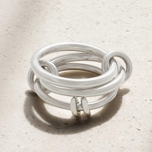 May include: A polished silver ring composed of multiple interlocking circular bands of varying thicknesses. The metal surface is smooth and reflective, featuring a small engraved SK mark on the interior of one of the bands. The design creates a layered, continuous loop structure.
