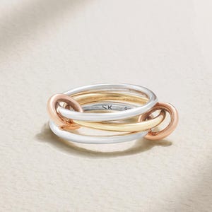 May include: A tri-color ring featuring three interlocking bands in silver, yellow gold, and rose gold tones. The silver band is engraved with the letters SK and the number 925 on the inner surface. The bands are smooth, polished, and circular.