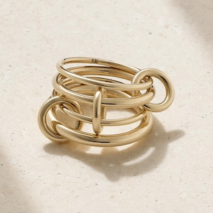 Può includere: Un anello in oro lucido caratterizzato da un design multistrato di fasce circolari interconnesse. La struttura è composta da diversi anelli d'oro lisci e arrotondati collegati tra loro in una disposizione complessa. Le lettere SK sono incise.