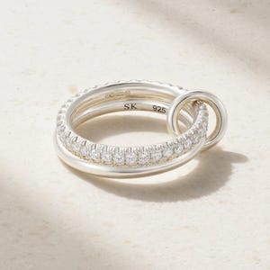 Könnte beinhalten: Ein Ring aus Sterlingsilber, bestehend aus einer glatten Ringschiene, die mit einem zweiten Ring verbunden ist, welcher mit einer Reihe klarer, rund geschliffener Steine besetzt ist. Innen sind die Buchstaben SK und die Zahl 925 eingraviert.