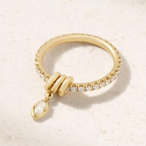 Op de afbeelding: Een gouden ring met een band bezet met kleine ronde witte stenen. De ring heeft een decoratief gedeelte met drie gouden lussen en een enkele witte steen in marquise-slijpvorm die aan het midden van de lussen hangt.