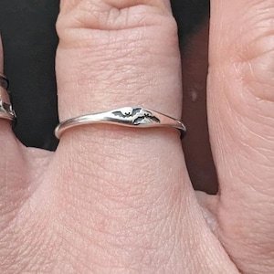 May include: A thin silver ring featuring a small, engraved bat silhouette centered on the band. The metal has a polished finish, and the bat design is highlighted with dark oxidation to define the wings and body shape clearly.