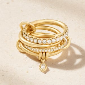 Può includere: Un anello in oro con molteplici fasce intrecciate, di cui due incastonate con piccole pietre bianche rotonde e una singola pietra taglio marquise che pende dal basso. Il design è composto da anelli in oro giallo lucido.