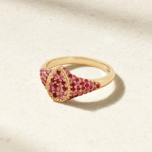 Könnte beinhalten: Ein goldener Siegelring mit einem tropfenförmigen Kopf, der mit kleinen, leuchtend rosa und roten Edelsteinen besetzt ist. Die Ringschiene ist glatt und poliert, während die Fassung um die Steine herum ein strukturiertes Detail aufweist.
