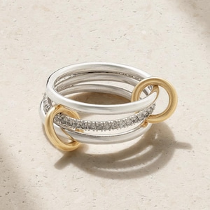 Può includere: Un anello a più fasce composto da tre fasce color argento e una fascia centrale incastonata con piccole pietre chiare. Due maglie circolari lisce color oro collegano le fasce sui lati, creando un design stratificato e interconnesso.
