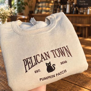 Könnte beinhalten: Ein beigefarbenes Sweatshirt mit Rundhalsausschnitt und dunkelbrauner Stickerei mit der Aufschrift PELICAN TOWN, EST. 2016 und PUMPKIN PATCH. Ein kleines, schwarzes, verpixeltes Katzensymbol ist mittig zwischen den Texten eingestickt.