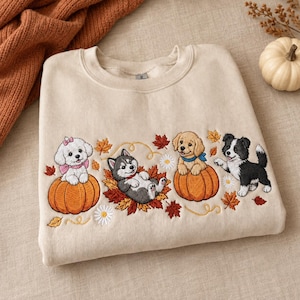 May include: A beige crewneck sweatshirt featuring an embroidered design of four puppies playing with pumpkins and autumn leaves. The embroidery includes a white puppy with a pink bow, a husky, a golden retriever, and a black and white puppy.