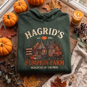 Könnte beinhalten: Ein dunkelgrüner Kapuzenpullover mit einem Grafikdruck und dem Text HAGRID'S PUMPKIN FARM EST. 1981 MAGICALLY GROWN. Das Design zeigt ein Steinhäuschen, eine schwarze Katze, ein Holzschild und einen Karren voller orangefarbener Kürbisse.