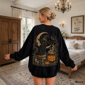 Könnte beinhalten: Ein schwarzes Langarm-Sweatshirt mit einem Grafikdruck auf der Rückseite. Das Motiv zeigt eine schwarze Katze mit Hexenhut, die auf einem Stapel Bücher neben einem leuchtenden Kürbis, Kerzen und einer Mondsichel in einem Wald sitzt.