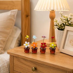Pode incluir: Quatro figuras de flores de vidro em miniatura em pequenos vasos castanhos. Cada flor apresenta pétalas de cores diferentes (branco, laranja, vermelho e amarelo) com uma pequena criatura de vidro no topo: uma abelha, um pássaro azul, uma borboleta e um pássaro vermelho.