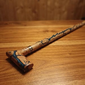 Könnte beinhalten: Ein Gehstock aus Holz mit T-förmigem Griff und langem Schaft, beide verziert mit komplexen, dunkelblauen blitzartigen Mustern, die in die Holzmaserung eingelassen sind. Ein silberfarbener Metallring mit Gravur befindet sich unter dem Griff.