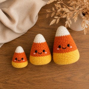 May include: Three crochet candy corn figures in varying sizes, each featuring a white top, orange middle, and yellow base. The figures have black bead eyes, a stitched black smile, and small pink embroidered cheeks on their textured surfaces.