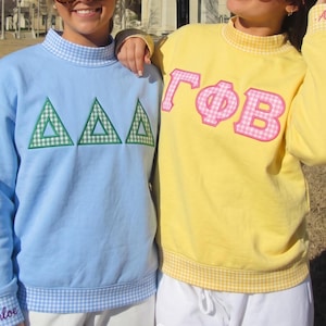 May include: A light blue sweatshirt with a mock neck and cuffs featuring a blue and white gingham pattern, displaying three green and white gingham Greek letters. A yellow sweatshirt with a mock neck and cuffs in yellow gingham, featuring pink and white gingham Greek letters and the name Anny embroidered on the cuff.