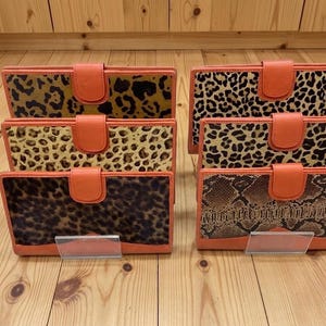 May include: Six rectangular wallets arranged in two rows of three, each featuring a solid orange leather trim and a snap-button closure. The wallets display various animal print patterns, including leopard spots and a brown snakeskin texture.