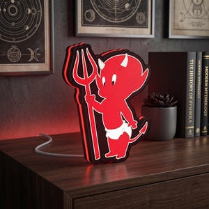 Puede incluir: Una luz LED roja estilo neón con forma de personaje de diablo de dibujos animados que lleva un pañal blanco y sostiene un tridente. La luz tiene un contorno negro, emite un brillo rojo intenso y tiene un cable de alimentación blanco.