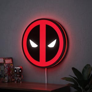 Può includere: Una lampada da parete a LED circolare con il logo Deadpool nero e rosso e occhi bianchi luminosi. L'insegna presenta un bordo circolare rosso vibrante e una striscia verticale rossa centrale su sfondo nero, emettendo una luce rossa brillante.