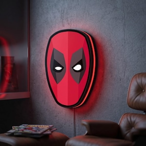 Può includere: Una lampada a LED da parete a forma di maschera di Deadpool. La maschera presenta un volto rosso vivace con contorni neri intorno agli occhi e occhi bianchi luminosi. La luce emette un alone rosso sulla parete e ha un cavo di alimentazione.