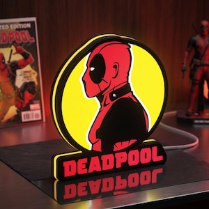 Puede incluir: Un letrero luminoso LED circular que presenta una ilustración de perfil roja y negra del personaje Deadpool sobre un fondo amarillo brillante. La base del letrero es negra con la palabra DEADPOOL impresa en letras mayúsculas rojas.