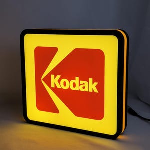 Może przedstawiać: Prostokątny, podświetlany szyld Kodak z jasnym żółtym tłem, wyrazistym czerwonym elementem graficznym i słowem Kodak wydrukowanym na żółto. Szyld posiada czarną ramkę i emituje ciepłe światło z krawędzi oraz przedniej powierzchni.