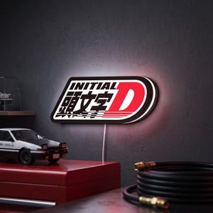 Könnte beinhalten: Ein rechteckiges LED-Wandschild mit dem Initial D-Logo, bestehend aus schwarzem, weißem und rotem Text sowie Grafiken. Das Schild zeigt das Wort INITIAL, japanische Schriftzeichen und ein großes rotes D, das ein sanftes weißes Licht ausstrahlt.