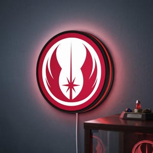 May include: A circular wall-mounted LED light featuring the white Jedi Order symbol centred on a vibrant red background. The sign has a thin black frame and emits a soft red glow from behind its perimeter, with a white power cord extending downwards.
