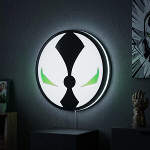 May include: A circular wall-mounted LED light featuring the black and white Spawn logo design with two sharp, glowing green accents. The light emits a soft white backlight around its perimeter, creating a halo effect against the dark wall surface.
