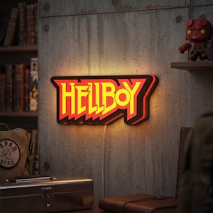 May include: A rectangular wall-mounted LED sign featuring the word HELLBOY in bold yellow lettering with a red outline and a black shadow effect. The sign emits a warm glow from behind its edges and includes a white power cord extending downwards.