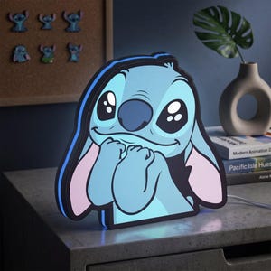 May include: A light blue Stitch character lamp with large dark eyes, a small nose, and long ears with pink inner sections. The character is depicted with hands held near the face in a shy pose, outlined in black with a glowing blue edge light.