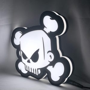 Peut inclure: Une enseigne murale lumineuse noire et blanche en forme de crâne et de tibias croisés stylisés. Le design présente un crâne aux allures de dessin animé avec des yeux triangulaires pointus et un sourire denté, souligné de noir.