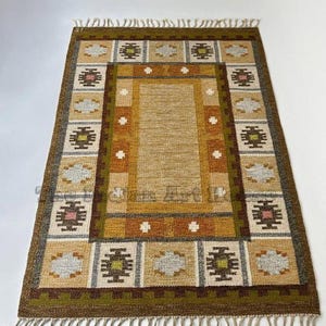 May include: A rectangular woven rug featuring a central tan panel surrounded by a border of geometric patterns in shades of brown, olive green, cream, and gray. The rug has a fringed edge on the top and bottom and a repeating motif design.
