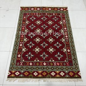 Puede incluir: Una alfombra rectangular con un centro rojo intenso y un patrón repetitivo de motivos de cruces geométricas en blanco y gris oscuro. La alfombra tiene un borde ancho verde salvia con diseños complejos de diamantes y cruces, con flecos blancos.