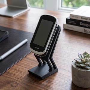 Könnte beinhalten: Ein weiß-schwarzer Garmin GPS-Fahrradcomputer auf einem schwarzen, abgewinkelten 3D-gedruckten Kunststoffständer. Das Gerät verfügt über einen rechteckigen Bildschirm mit schwarzem Rahmen und dem Garmin-Logo in Weiß unten in der Mitte.